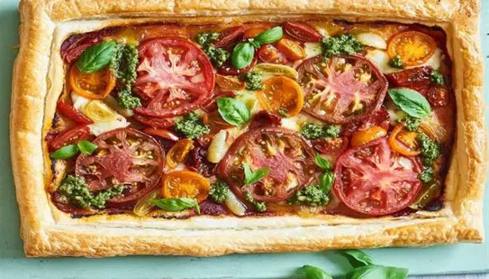 A golden-brown puff pastry tart topped with sliced tomatoes, sun-dried tomatoes, melted Gouda cheese, and fresh basil leaves.